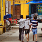 Three boys walking arm in arm with their backs facing the camera in a street in India.