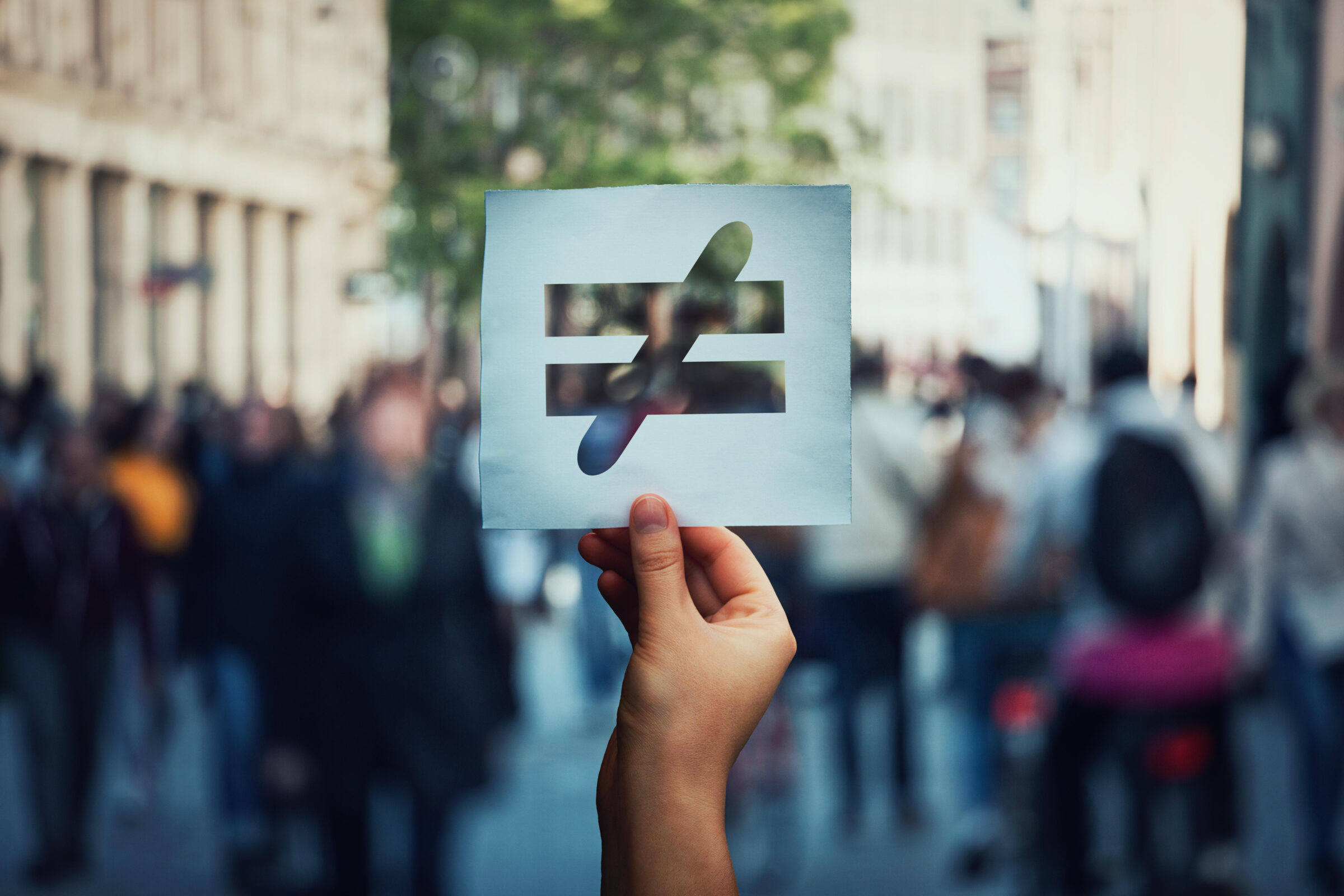 unequal sign cut into paper held in a crowd with a blurred background