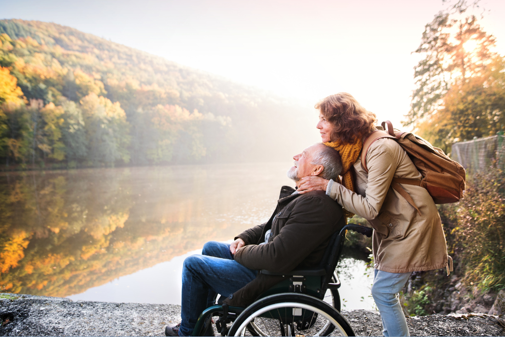 Baby boomer woman with chestnut brown shoulder length hair wearing a tan jacket and brown backpack stands over baby boomer man with short grey hair, wearing a dark jacket and sitting in a wheelchair. Both look out over a river to the forest on the other bank.