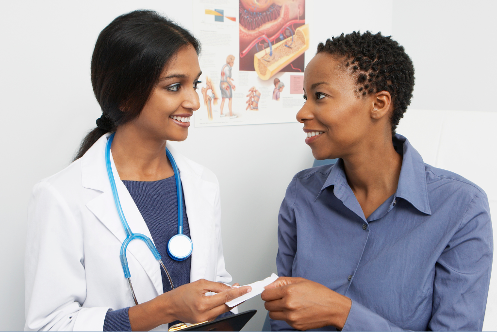 Doctor with long black hair in a ponytail and blue stethoscope around her neck gives patient information to a woman with short black hair wearing a blue button down shirt.