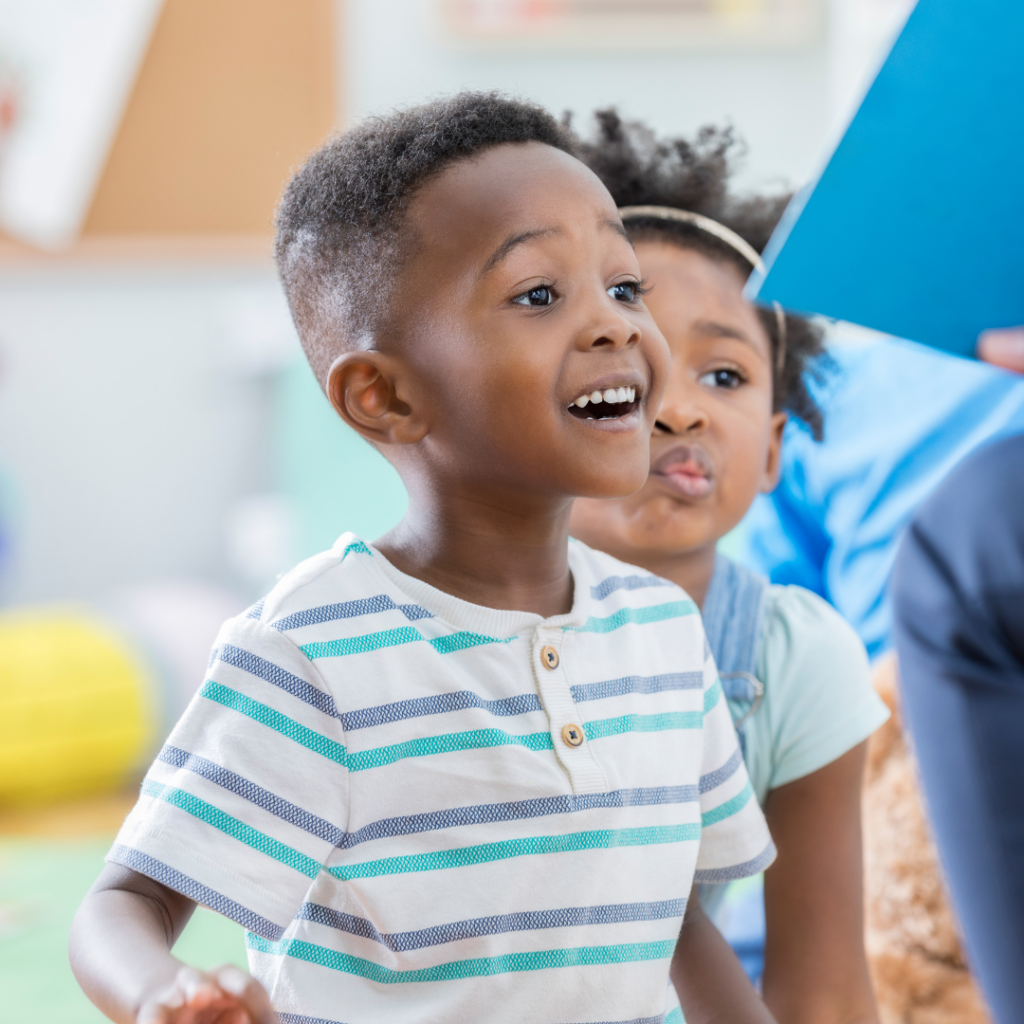 African American preschool boy wearing a white and blue striped shirt is excitedly looking at a book held by a teacher.