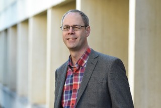 A portrait photo of Professor Anthony J. Langlois. The photo is from the mid-torso upwards, with the background indicating they are stood outside with a concrete building backdrop. Prof. Langlois is wearing glasses, a blue, red, and white plaid button shirt, and a warm grey blazer. They have white skin and light brown hair.