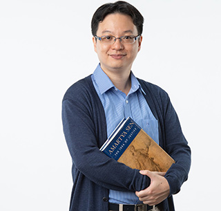 A standing portrait photo of Benedict S. B. Chan, an Associate Dean and Associate Professor at the Hong Kong Baptist University. B.S.B Chan has very light brown skin, and is wearing a light blue shirt with a dark blue cardigan on top, and light brown trousers with a brown belt. They are holding a book in their arms, titledL 'Amartya Sen: The Idea of Justice'. The background is a very light grey colour.
