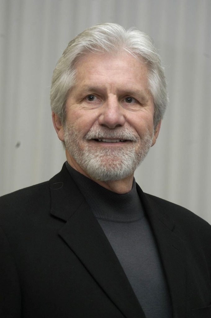 A portrait image of Terry O'Banion against a light and mid grey background. He has white hair, fair skin, a white moustache and white beard while wearing a dark grey high neck jumper and black blazer.
